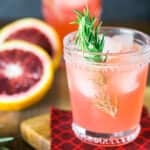 A blood orange gin and tonic on a wooden tray with fabric and fruit beside it.