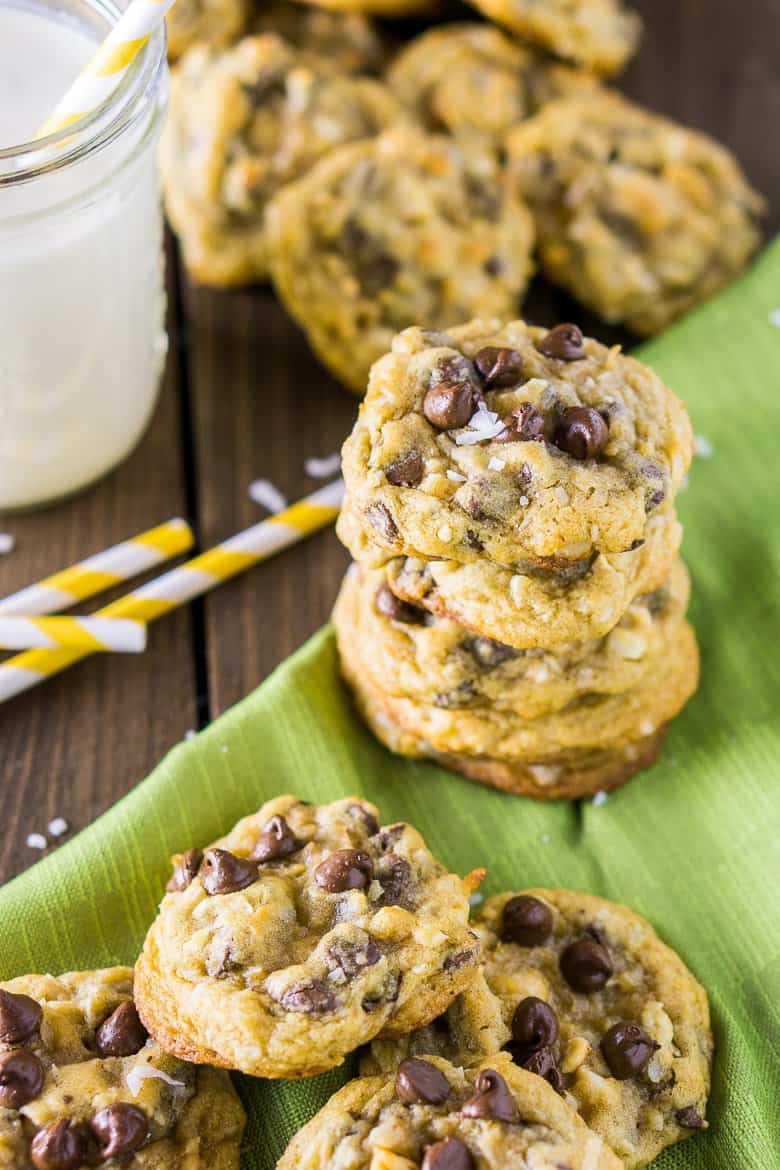 A stack of chocolate chip cookies with coconut and macadamia nuts on a green napkin with milk and straws next to it.