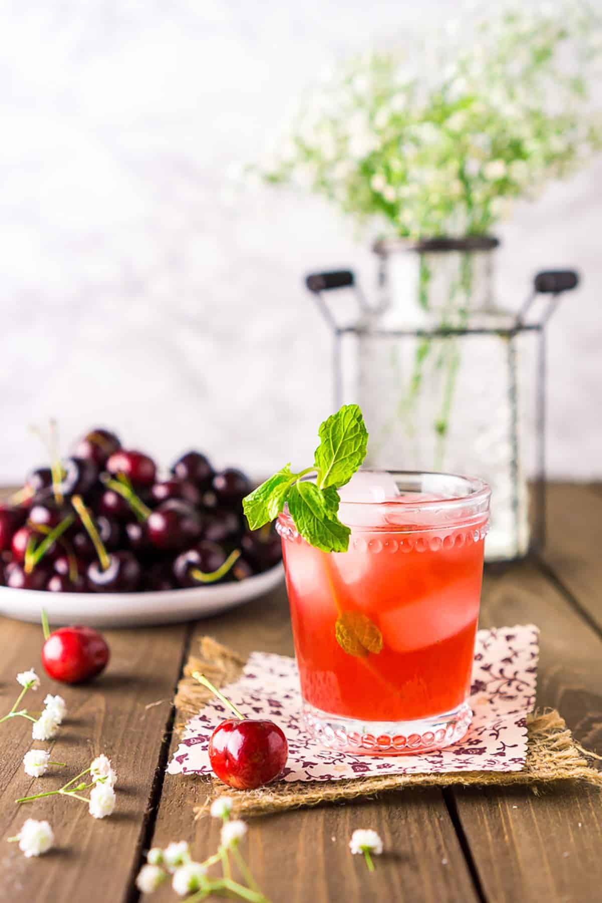 A cherry bourbon smash on burlap with a plate of cherries and white flowers in the background.