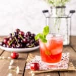 A cherry bourbon smash on burlap with a plate of cherries and white flowers in the background.
