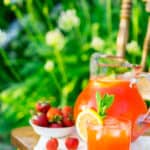 A pitcher of minted strawberry lemonade on a wooden chair in a garden.