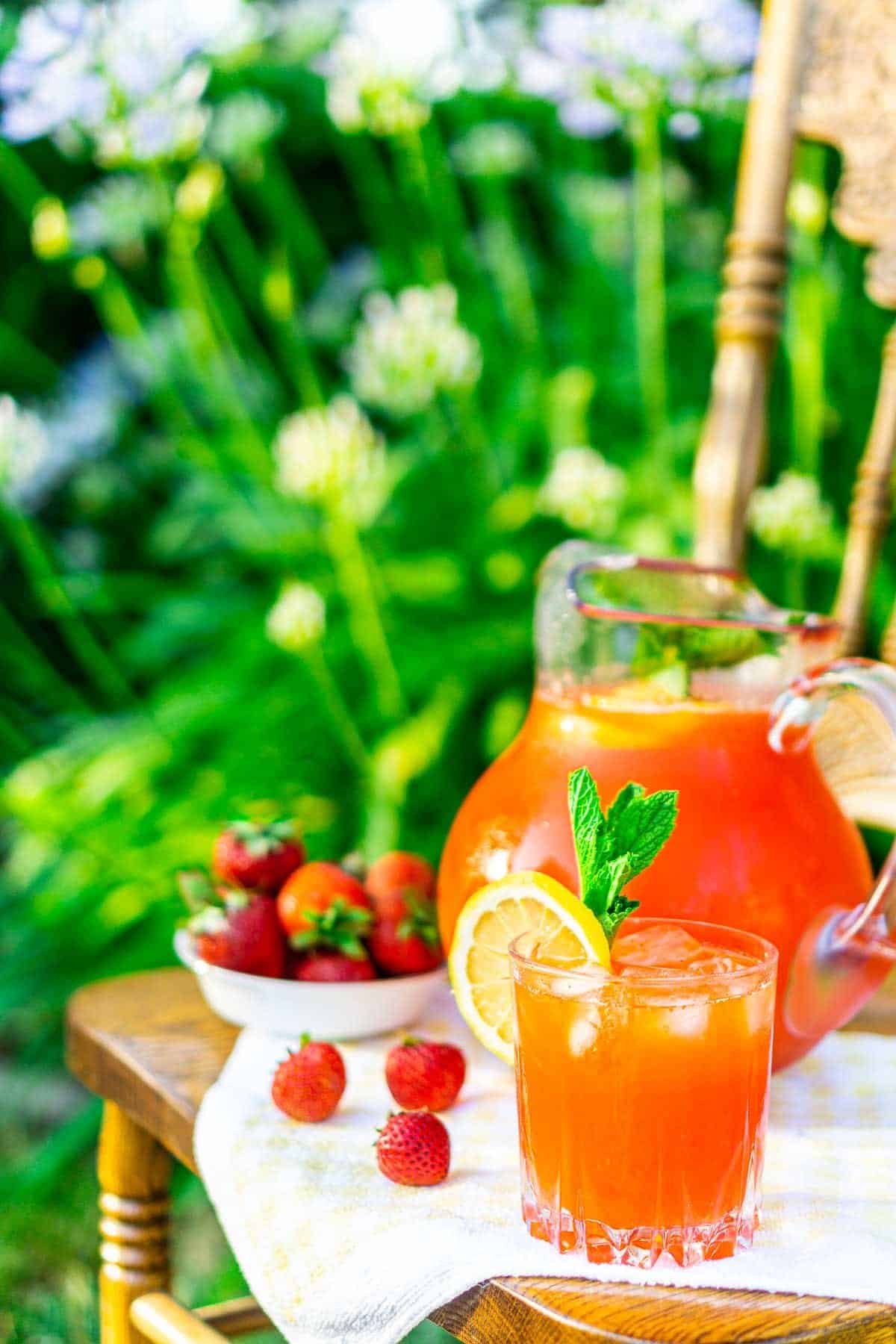 A glass of strawberrry-mint lemonade on a chair in a garden with a pitcher behind it.