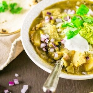 A bowl of Colorado pork green chili on a brown painted surface with a spoon in it and red onions and tortillas to the left.