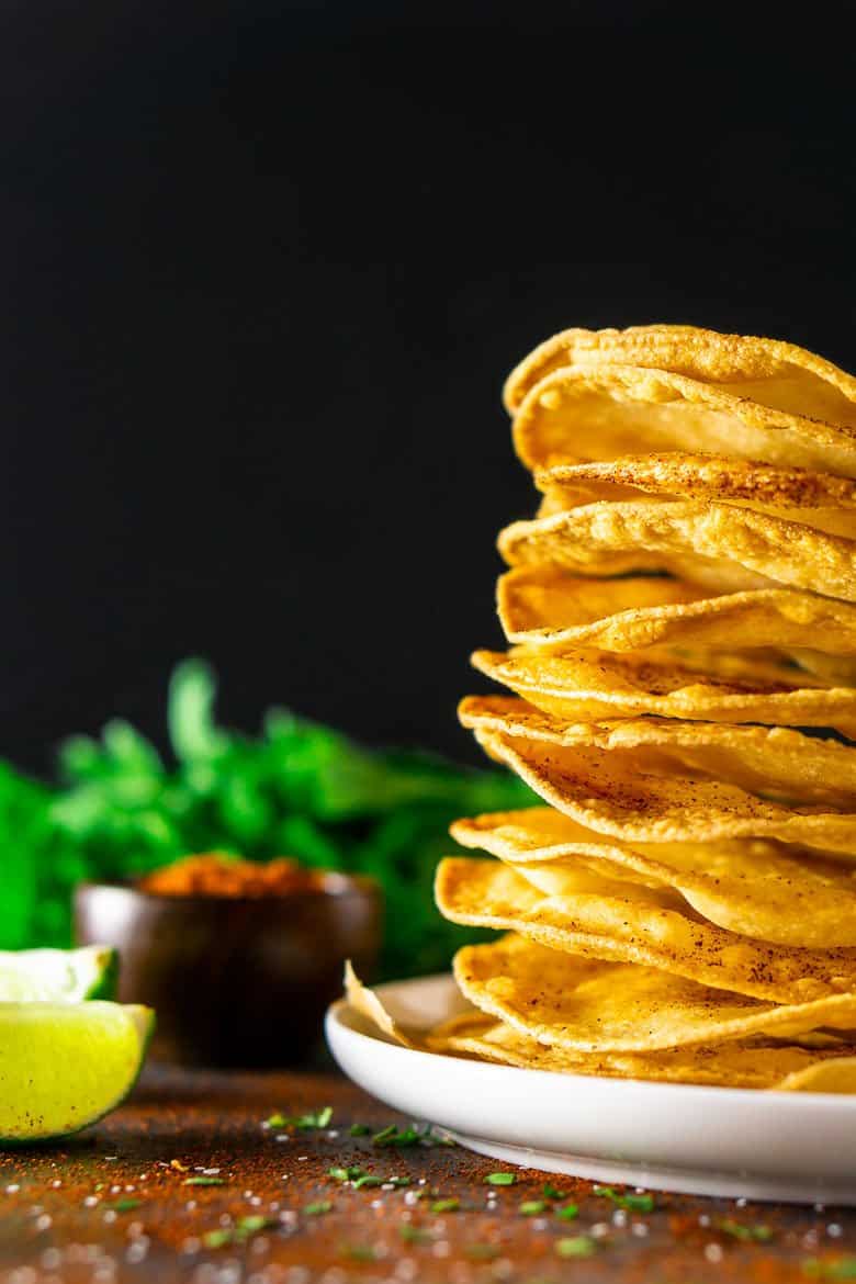 Freshly fried tostada shells stacked against a black background with cilantro, chili powder, lime and salt.