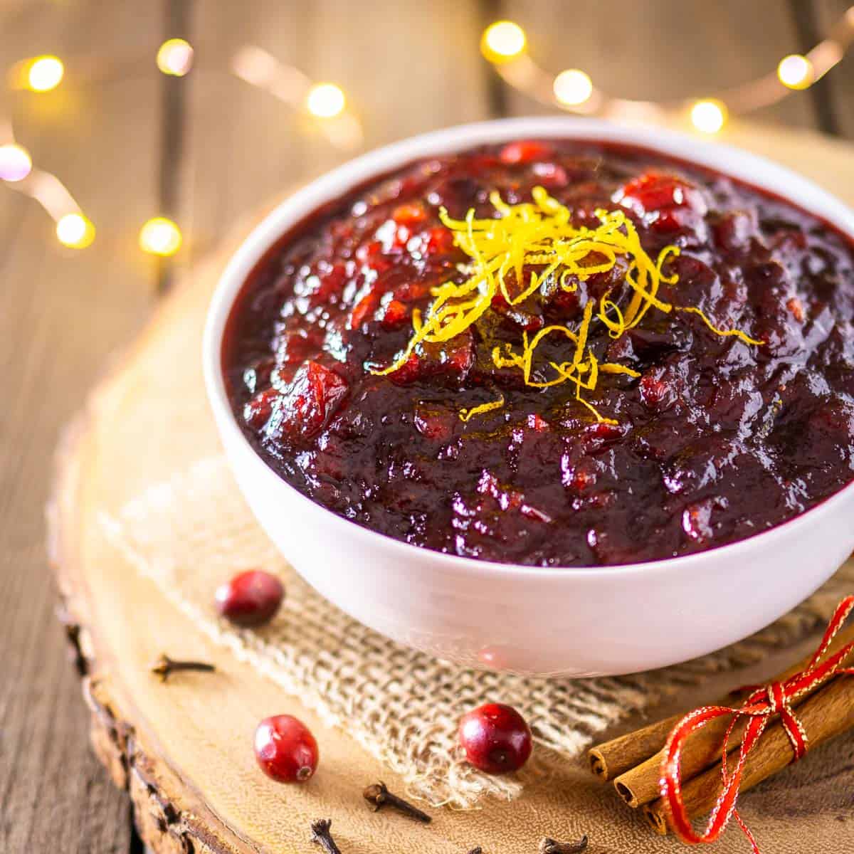 A bowl of brandy cranberry sauce on a wooden platter with twinkle lights behind it.
