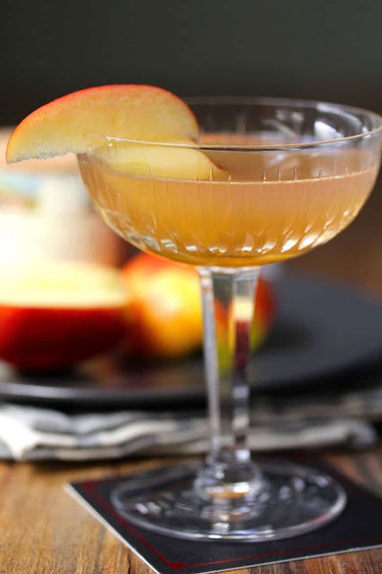 An Apple Brown Derby Cocktail with apple slices in the background. 