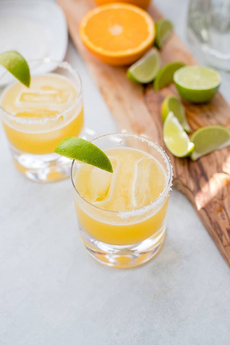 Two skinny margaritas with a wooden board holding limes and oranges.