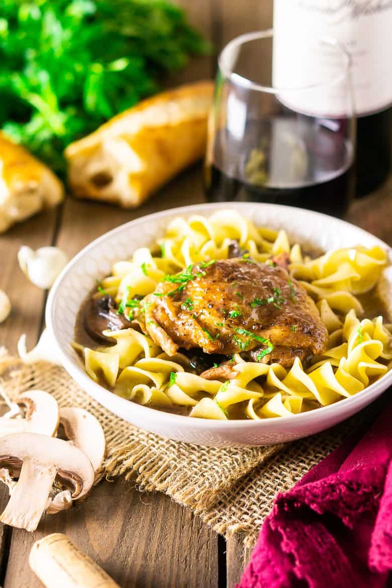 A bowl of coq au vin with bread, mushrooms, a wine cork and glass of wine.