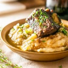 A bowl of brown butter mashed potatoes with a red wine-braised short rib on top with a glass of wine.