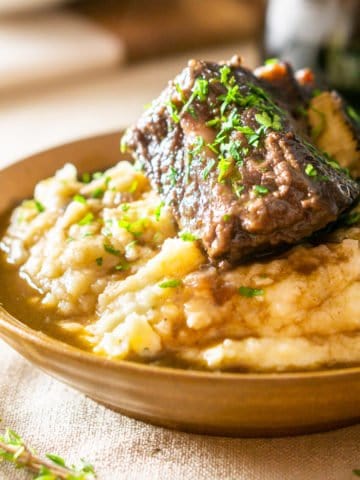 A bowl of brown butter mashed potatoes with a red wine-braised short rib on top with a glass of wine.