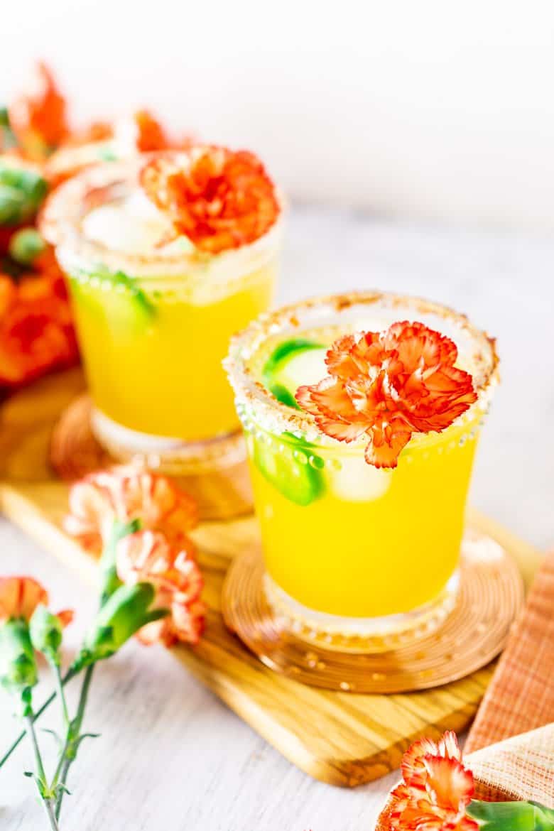 Two jalapeno-mango margaritas on a wooden board with flowers.