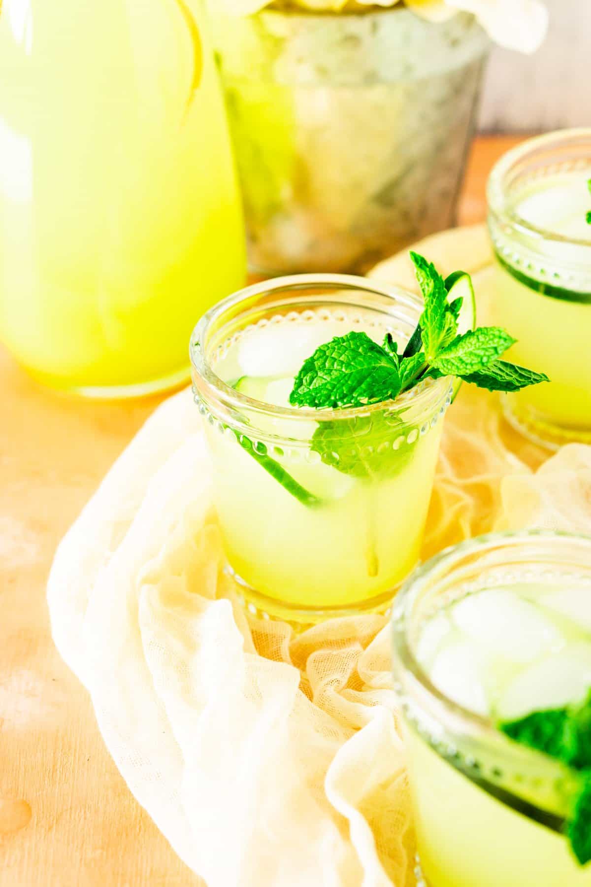 A glass of mint-cucumber lemonade with two other glasses in the foreground and background.