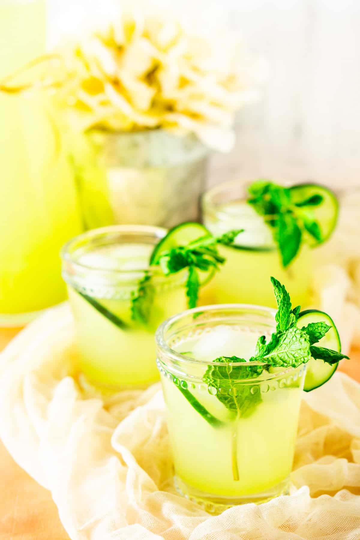 Three glasses of mint-cucumber lemonade with flowers and a carafe.