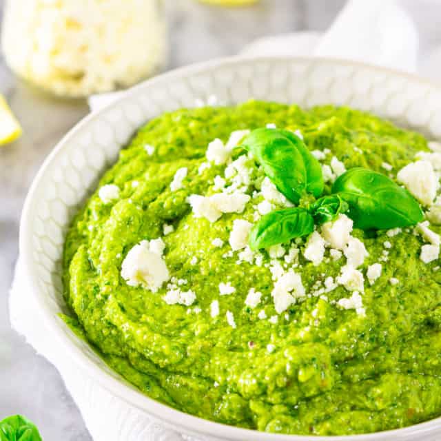 A bowl of feta-avocado pesto sauce with a jar of feta and lemon slices in the background.