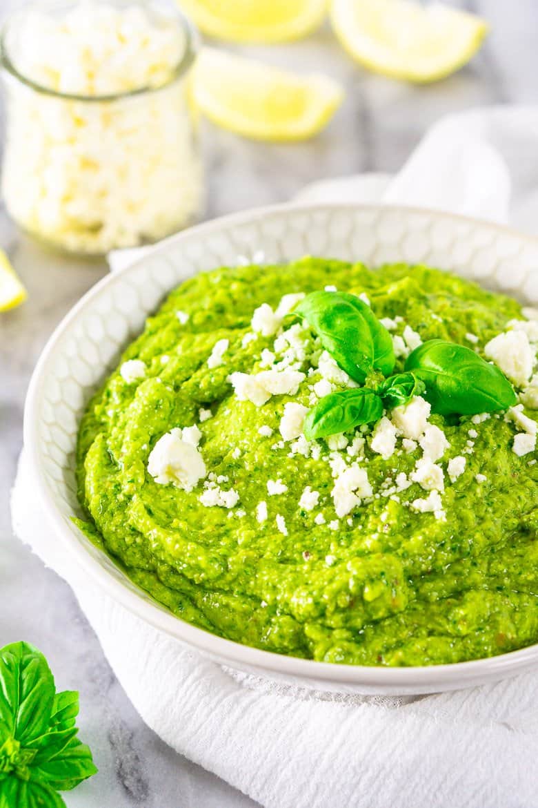 A bowl of feta-avocado pesto sauce with a jar of feta and lemon slices in the background.