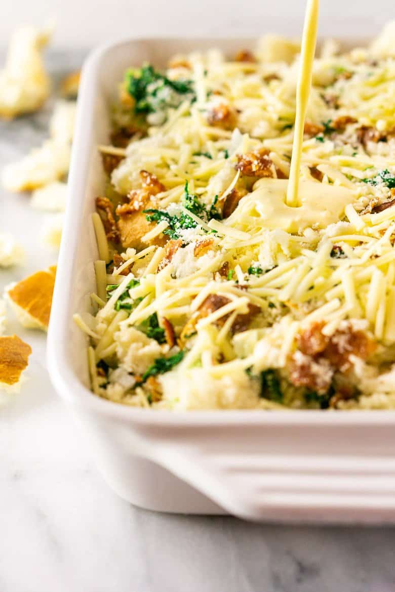 Pouring the egg mixture over bacon, Gruyere and spinach strata in a white pan.