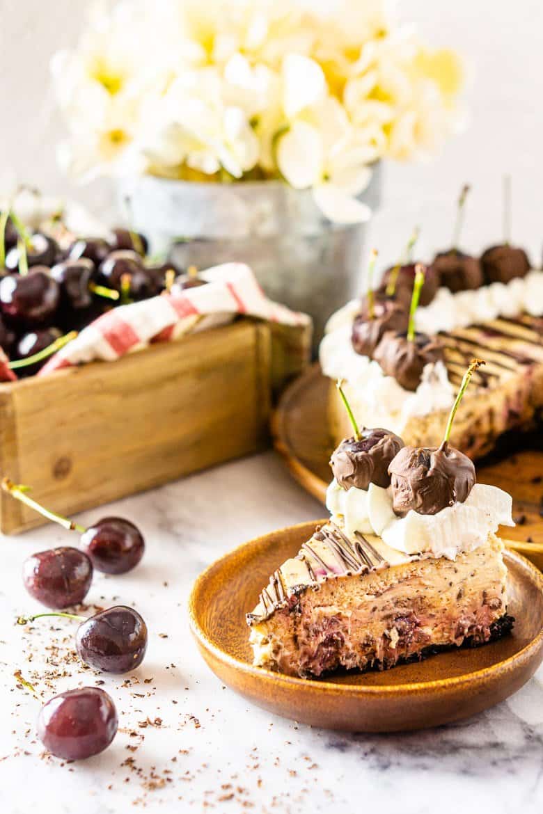 A slice of chocolate-covered cherry cheesecake on a wooden plate with the full cheesecake in the background with more cherries and flowers.