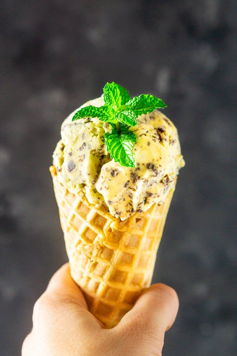 A hand holding a waffle cone with fresh mint-chocolate chip ice cream against a black and gray wall.