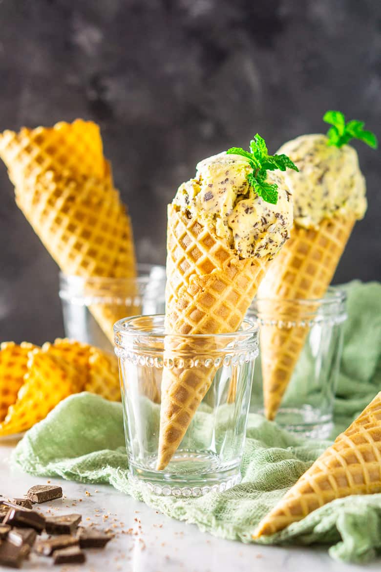 Two waffle cones filled with fresh mint-chocolate chip ice cream with chopped chocolate and empty waffle cones in the background.