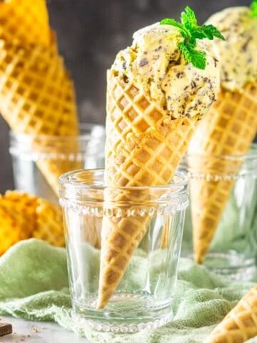Two waffle cones filled with fresh mint-chocolate chip ice cream with chopped chocolate and empty waffle cones in the background.