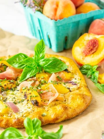 A grilled peach pizza on parchment paper with fresh peaches and purple flowers.