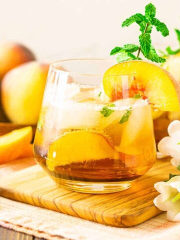 A peach bourbon smash on a wooden tray with a bunch of white flowers.