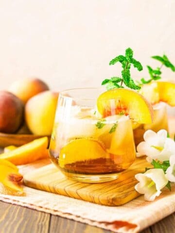 A peach bourbon smash on a wooden tray with a bunch of white flowers.