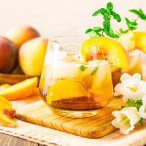 A peach bourbon smash on a wooden tray with a bunch of white flowers.