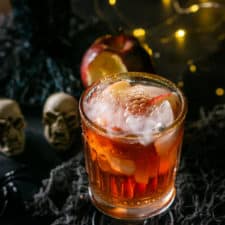 A single Poisoned Orchard Cocktail with steam coming toward you with lights in the background.
