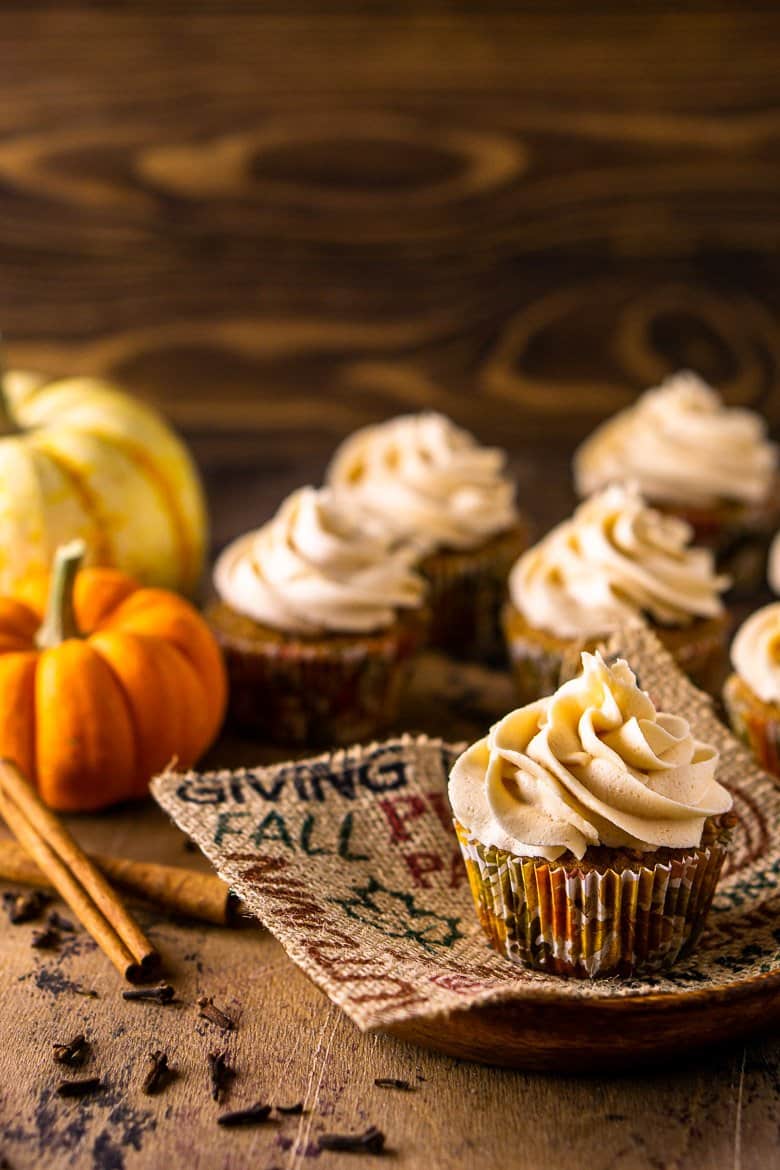 A brown butter pumpkin cupcake on a fall-themed piece of burlap with pumpkins, cinnamon sticks and other cupcakes in the background.