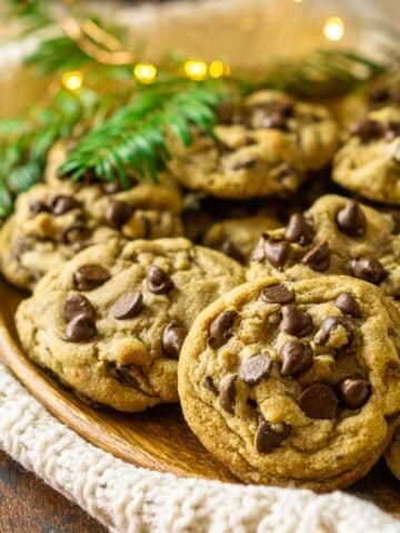 A plate of spiced holiday chocolate chip cookies on a wooden plate with pine needles and lights in the background.