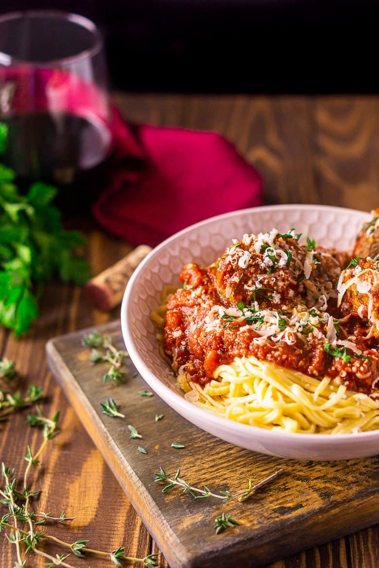 A bowl of Italian meatballs and spaghetti with a glass of wine and burgundy napkin in the background along with fresh herbs.