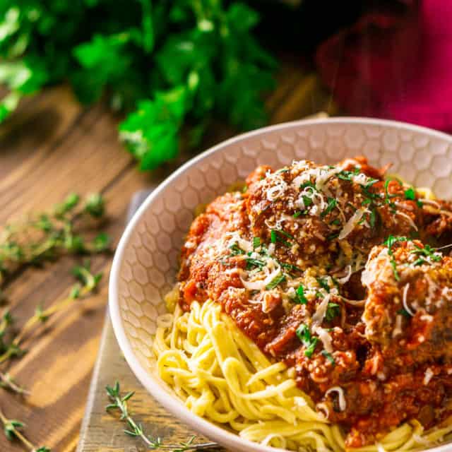 A bowl of Italian meatballs and spaghetti on a wooden tray with fresh parley, thyme and wine in the background.