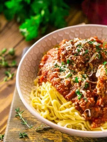 A bowl of Italian meatballs and spaghetti on a wooden tray with fresh parley, thyme and wine in the background.