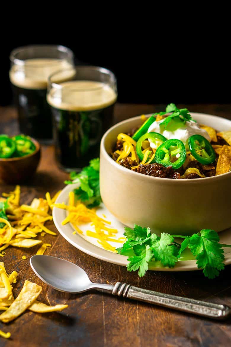 A bowl of authentic Texas chili with cilantro, cheese and two stouts in the background.
