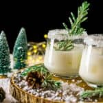 A Winter Wonderland Margarita on a wood board with candied rosemary and fake trees and lights in the background.