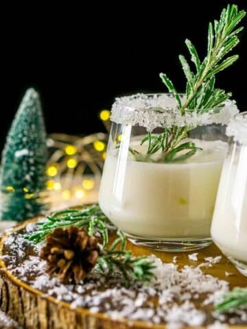 A Winter Wonderland Margarita on a wood board with candied rosemary and fake trees and lights in the background.