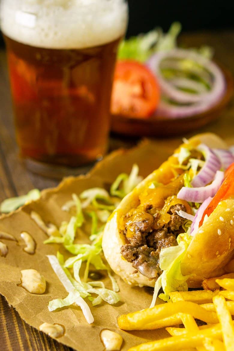 A cheeseburger sub on brown paper with fries on the side and a beer with a plate of toppings.