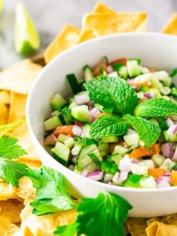 A bowl of cucumber pico de gallo with pita chips and herbs around it.