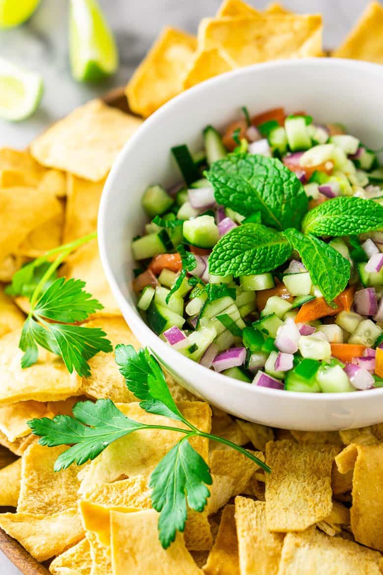Looking from the side to a bowl of cucumber pico de gallo with herbs, chips and limes.