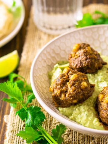 Mexican meatballs in a shallow bowl with avocado-crema sauce with cilantro and lime slices.