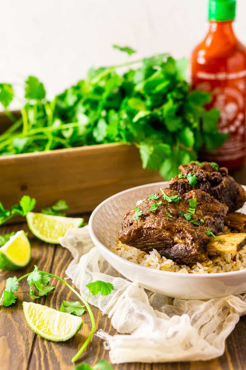 Two braised sriracha short ribs over coconut rice with cilantro, limes and a bottle of sriracha in the background.