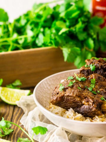Two braised sriracha short ribs over coconut rice with cilantro, limes and a bottle of sriracha in the background.