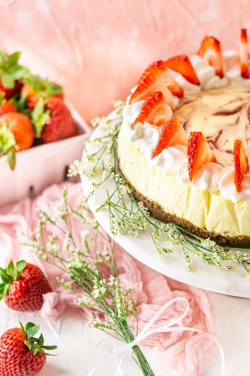 The fresh strawberry-ginger cheesecake on a white marble cake stand with white flowers around it.