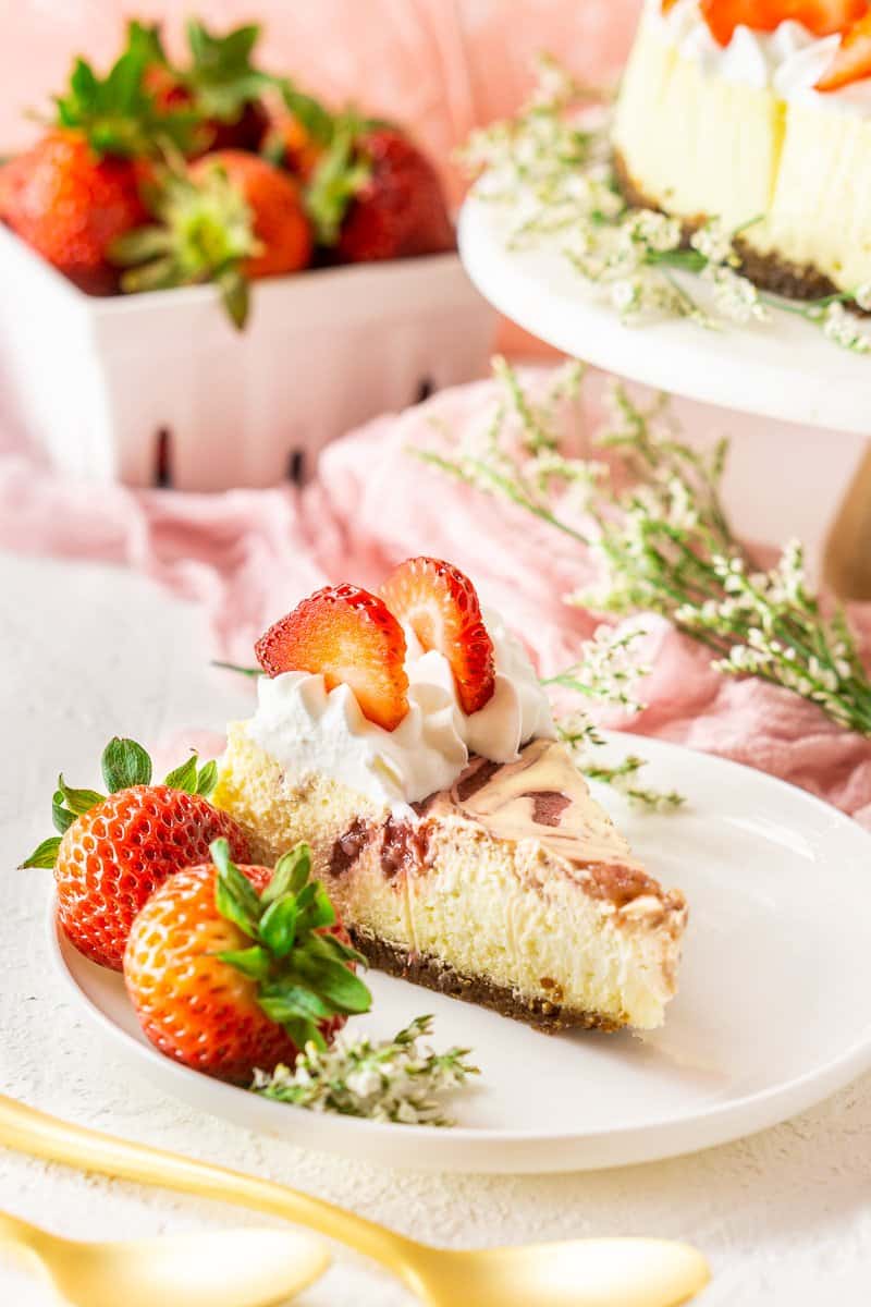 A slice of the fresh strawberry-ginger cheesecake with strawberries, flowers and the whole cheesecake in the background.