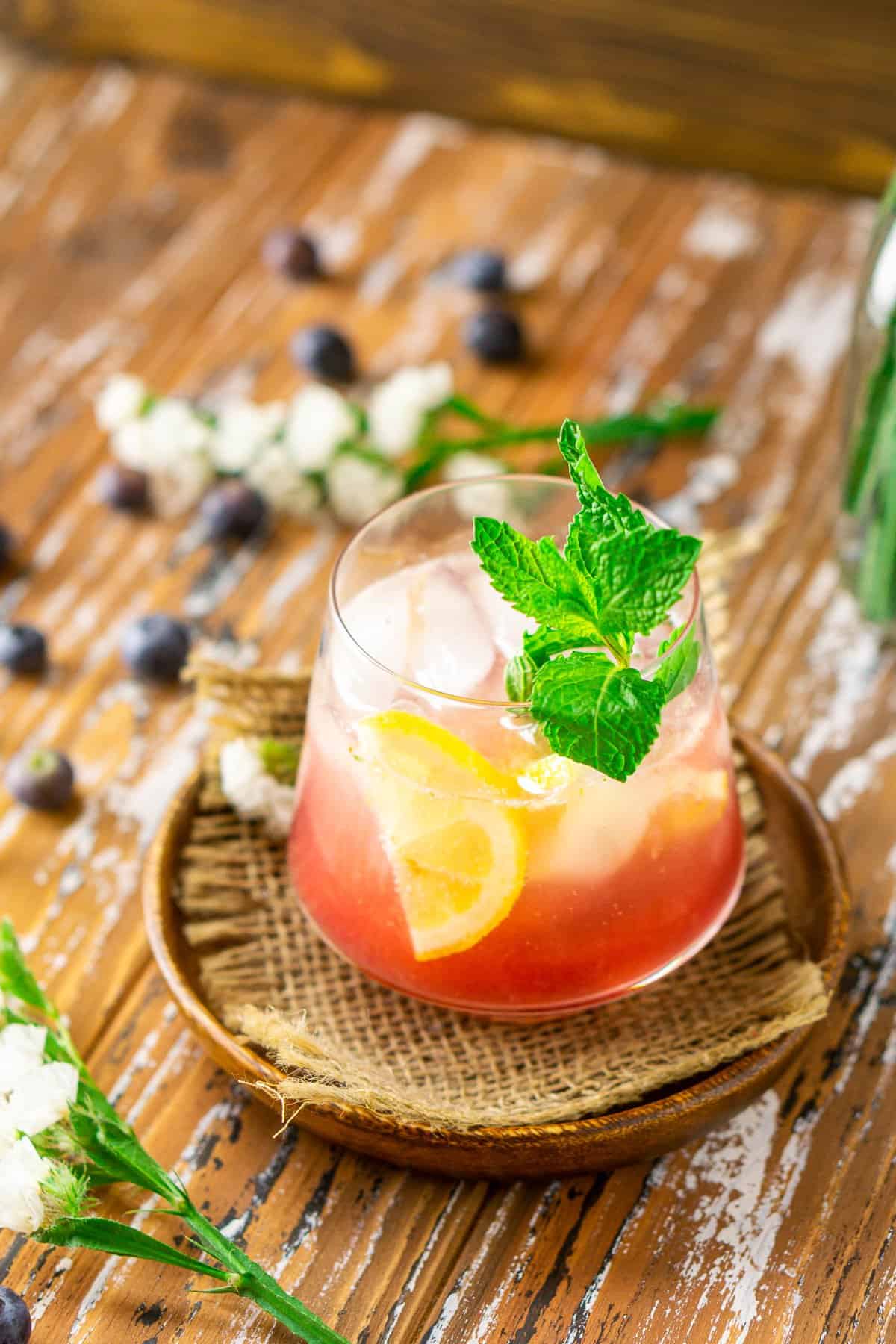 A side view of the fresh blueberry cocktail with flowers and blueberries surrounding it.