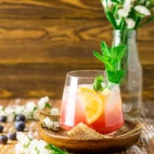 A blueberry bourbon smash on a wooden plate with white flowers in the background.