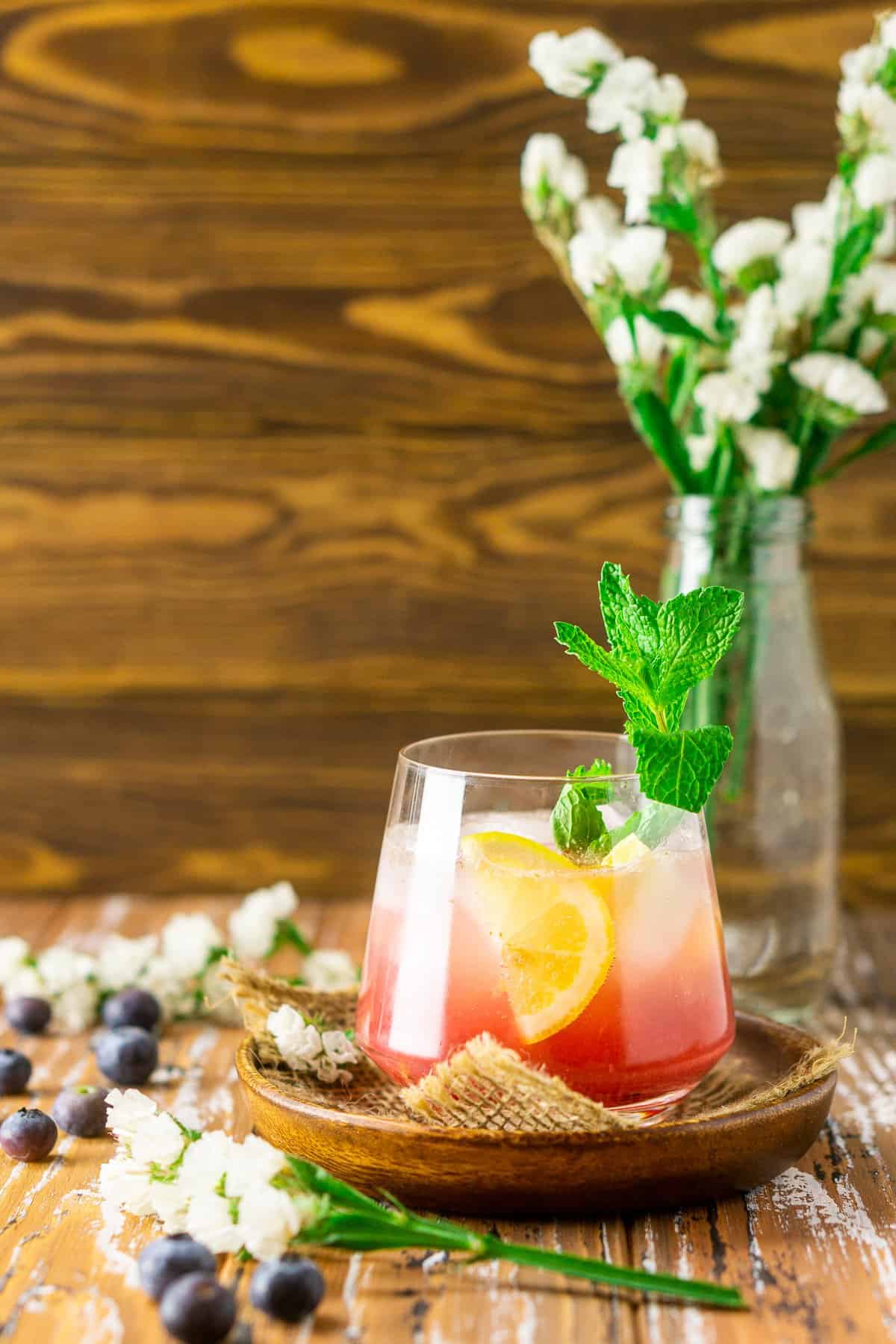 A blueberry bourbon smash on a wooden plate with white flowers in the background.