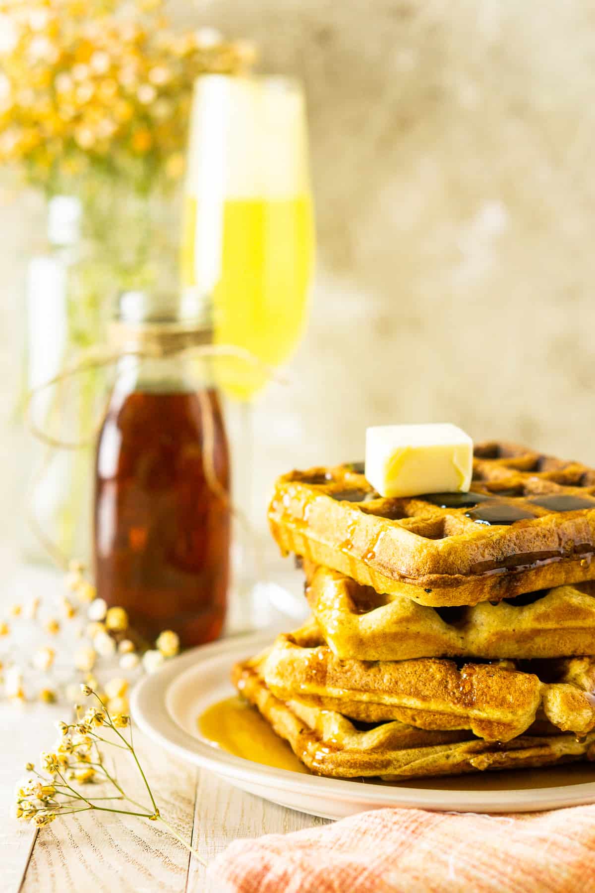 A stack of perfect buttermilk waffles with baby's breath flowers, syrup and a mimosa.