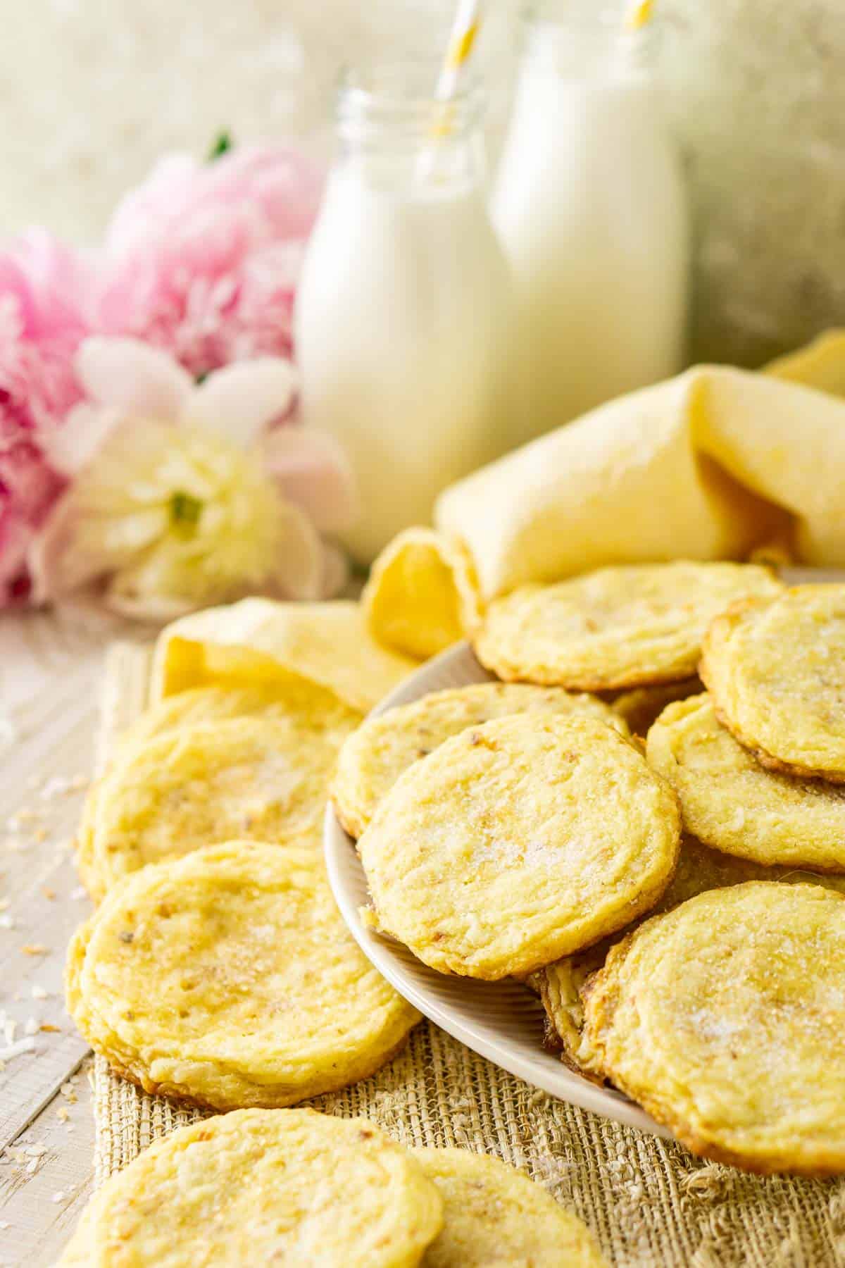 A plate of toasted coconut sugar cookies with coconut on the side and flowers and milk in the background.